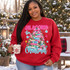 All Booked for Christmas DTF transfer featuring a Christmas tree made of colorful patterned books with pine branches, string lights, pinecones, and a pink plaid bow Shown on a Cardinal Red crewneck sweatshirt.