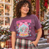 Christmastown snow globe DTF transfer with Christmas village, red truck, pink gingham bow, and pastel stars and snowflakes on a purple shirt Shown on a Dusk t-shirt.