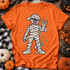 Rock On Mummy Halloween DTF Transfer | Funny Cartoon Mummy Design, playful bandaged cartoon mummy making a rock hand sign printed on an orange T-shirt with colorful splatters, surrounded by Halloween pumpkins and decor.