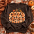 Cozy Season DTF Transfer | Gingham Plaid Fall Bubble Letter Design, orange gingham bubble letters spelling “Cozy Season” with a pumpkin stem and autumn leaves displayed on a brown sweatshirt among fall plaid fabric, pumpkins, and a cup of tea.
