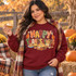 Happy Thanksgiving DTF Transfer | Hand-Painted Turkey & Holiday Feast Design, woman wearing a burgundy sweatshirt with a colorful Thanksgiving turkey and holiday feast design at an autumn pumpkin patch