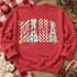 Christmas Mama DTF transfer with chunky block letters filled in candy-cane stripe, holly, checkerboard, and leopard patterns, topped with red cursive mama script and holiday bow, bell, and candy cane accents Shown on a Heather Scarlet Red crewneck sweatshirt.