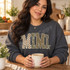 MIMI block letter DTF transfer with checkerboard, camo, leopard, and polka dot patterns, cream outlines, and ivory ribbon bows on an olive shirt Shown on a Dark Heather crewneck sweatshirt.