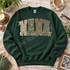 NANA block-letter DTF transfer in checkerboard, camo, leopard, and polka dot patterns with cream ribbon bow accents Shown on a Forest crewneck sweatshirt.