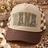 NANA block-letter DTF transfer in checkerboard, camo, leopard, and polka dot patterns with cream ribbon bow accents Shown on a Khaki front panel with brown bill and brown-and-cream checkered mesh back trucker hat.