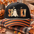 Spooky folk-art DTF transfer with patterned patchwork letters, a ghost replacing the O, a jack-o-lantern by the K, and flying bats above Shown on a black foam front panel with an orange-and-black striped bill and cream rope, black mesh back trucker hat.