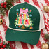 Merry Christmas DTF transfer featuring a flocked white Christmas tree with a pink gingham bow, yellow star, and colorful hand-lettered Merry Christmas text on a red polka-dot striped background with snowflake and candy cane stickers Shown on a pine green front panel, cream rope, white mesh back trucker hat.