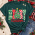 Merry Christmas DTF transfer with patterned block letters, a decorated Christmas tree with gold star and pink bows, and red script Christmas lettering Shown on a Blue Spruce t-shirt.