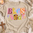 Blessed DTF transfer with scrapbook-style polka-dot, plaid, and star paper cutout letters and a pink ribbon bow Shown on a Sandstone t-shirt.