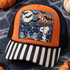 Halloween DTF transfer featuring a cartoon beagle holding a BOO! sign in a starry graveyard scene with a full moon, bats, tombstones, skulls, bones, and pumpkins Shown on a burnt orange front panel with black and white striped bill and black mesh back trucker hat.