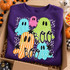BOO Halloween DTF transfer featuring five patterned cartoon ghosts in teal, pink, green, and orange on a black and white speckled background with stars Shown on a Purple crewneck sweatshirt.