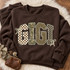 GIGI DTF transfer in oversized varsity letters with checkerboard, camo, leopard, and polka dot patterns and cream bow accents Shown on a Dark Chocolate crewneck sweatshirt.