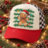 Gingerbread man DTF transfer wearing a sparkly Santa hat and plaid bow, surrounded by pine greenery and colorful stars Shown on a cream front panel with red bill and black-and-white checkered mesh trucker hat.