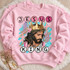 Jesus is King DTF transfer with painterly portrait of Jesus wearing a golden crown over a crown of thorns, teal brushstrokes, pink florals, and circle-letter text Shown on a Light Pink crewneck sweatshirt.