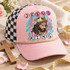 Jesus is King DTF transfer with painterly portrait of Jesus wearing a golden crown over a crown of thorns, teal brushstrokes, pink florals, and circle-letter text Shown on a Blush front panel, gold rope, black-and-white checkered mesh back trucker hat.