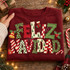 Feliz Navidad DTF transfer with oversized mixed-pattern lettering in leopard print, checkerboard, stripes, dots, and painted Christmas trees, accented by a gold bell, holly berries, a candy cane, and a snowman ornament Shown on a Maroon crewneck sweatshirt.