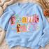 Thankful DTF transfer in whimsical torn-paper collage lettering with mismatched patterns, pink ribbon bow, and hand-drawn stars Shown on a Light Blue crewneck sweatshirt.