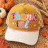 Thankful DTF transfer in whimsical torn-paper collage lettering with mismatched patterns, pink ribbon bow, and hand-drawn stars Shown on a mustard gold front panel with cream bill and cream-and-mustard checkered mesh back, pink rope trucker hat.