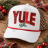 Yule-ish Christmas DTF transfer with mixed buffalo plaid, leopard print, distressed, and checkerboard pattern lettering and dark green brush-script ish. text Shown on a white front panel with red rope and leopard mesh back trucker hat.