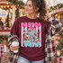 All Booked for Christmas DTF transfer with a whimsical book-stack Christmas tree, pine garland, fairy lights, and pink bubble lettering on a striped polka-dot background Shown on a Wine t-shirt.
