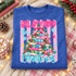 All Booked for Christmas DTF transfer with a whimsical book-stack Christmas tree, pine garland, fairy lights, and pink bubble lettering on a striped polka-dot background Shown on a Heather Deep Royal crewneck sweatshirt.