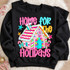 Home For The Holidays DTF transfer with colorful bubble lettering and a candy-coated gingerbread house on a pink polka-dot background Shown on a Black crewneck sweatshirt.