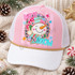 Let It Snow DTF transfer with a smiling patchwork snowman in a pink and teal striped hat and scarf, framed by a gold-glitter-style scalloped medallion with snowflakes and stars Shown on a Light Pink front panel, white bill, white mesh back trucker hat.