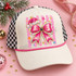 Pink Santa hat and glittery pink bow DTF transfer with black belt buckle center, stars, snowflakes, and polka-dot stripe background on a lavender shirt Shown on a ivory front panel with pink rope and black-and-white checkered mesh trucker hat.