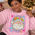 Whimsical Santa face DTF transfer with dotted fur trim, swirly beard, and patchwork hat inside a pink and teal scalloped frame Shown on a Light Pink crewneck sweatshirt.