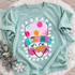 Colorful snowman face DTF transfer with polka-dot striped hat, carrot nose, pink cheeks, and winking eyes framed by a fringed border Shown on a Seafoam t-shirt.
