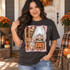 Trick or Moo Halloween DTF transfer featuring a highland cow in a ghost costume holding a glowing jack-o'-lantern bucket, with retro orange and brown striped background, bats, and stars Shown on a Pepper t-shirt.