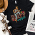 Dirty Cowgirl Screen Print Transfer, a black t-shirt featuring a cartoon cowgirl with the word 'Dirty' in leopard print and 'cowgirl' in blue script, surrounded by colorful flowers.