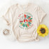 I Got A Peaceful Easy Feeling Screen Print Heat Transfer, a beige t-shirt with a colorful peace sign surrounded by feathers and peace symbols, laid out on a white surface with a sunflower and bracelets nearby.