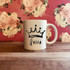 Queen Sublimation Transfer, white ceramic mug with a black crown and the word "Queen" printed on it, floral pink background.