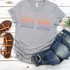Boss Babe Screen Print Heat Transfer, colorful graphic t-shirt with 'Boss Babe' text in pink, orange, purple, and yellow colors laid out on a white wooden surface with denim jeans and brown sandals nearby.