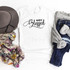 Simply Blessed Sublimation Transfer, a white T-shirt with black decorative text, placed among a gray hat, a floral scarf, and a pair of jeans with a gray knit sweater, showcasing vibrant clothing accessories.