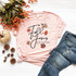 Fall for Jesus He Never Leaves Screen Print Transfer, pink T-shirt with autumn leaves and inspirational message on white wooden surface, with jeans, pumpkins, berries, and pinecones nearby.