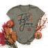 Fall for Jesus He Never Leaves Screen Print Transfer, gray T-shirt with fall leaves design and inspirational message, surrounded by pumpkins and autumn flowers for seasonal decoration