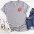 I Know I Play Like A Girl Softball POCKET/CAN HUGGER Screen Print Heat Transfer T-shirt, gray short-sleeve shirt with colorful softball graphic and motivational text, alongside denim jeans and white sneakers on a white background.