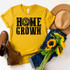 Home Grown Screen Print Heat Transfer, yellow t-shirt with black graphic design, surrounded by sunflowers, hat, denim jeans, and brown boots, stylish outdoor fashion apparel.