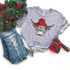 Shaggy Cow Hat Screen Print Heat Transfer, graphic t-shirt with a shaggy cow wearing a red and green striped Christmas hat with pom-pom, placed on a flat surface along with distressed jeans, a small holiday gift box, and Christmas decorations.