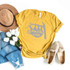 Drop It Like A Sloth FINAL STOCK Screen Print Heat Transfer, yellow T-shirt with a sloth illustration and humorous text, laid flat with casual accessories including jeans, white tulips, a straw hat, and beige slip-on shoes.