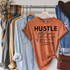 Hustle til the Haters Black Screen Print Heat Transfer T-shirt, orange t-shirt with black and cursive text, hung on a clothing rack with jeans, plaid blanket, and accessories.
