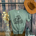 Life Update Still A Mess Print Heat Transfer, a gray T-shirt with black text and hashtag, displayed on a hanger with a frayed edge knot, hanging in front of a rustic wooden background, styled with a brown hat, a glass vase with artificial flowers, and sneakers.