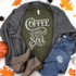 Coffee Warms The Soul Heat Transfer T-shirt, dark green with white text, surrounded by a gray knit cardigan, blue ripped jeans, beige ankle boots, and autumn leaves and pumpkins.