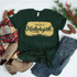Raise A Hallelujah Screen Print Heat Transfer, green t-shirt with 'Raise A Hallelujah' text design on it, surrounded by Christmas decorations including a plaid scarf, pine branches with red berries, pinecones, a pair of beige ankle boots, and jeans.