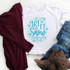 Let It Snow Somewhere Else Sublimation Transfer, white T-shirt with blue snowflake and festive text design, casual jeans, brown boots, and maroon sweater on white wooden surface.