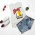 Busy Raising Ballers Baseball/Softball Screen Print Heat Transfer T-Shirt with graphic of a baseball with a red polka dot bow, paired with denim shorts, red sunglasses, white sneakers, and a green monstera leaf keychain on a white background.