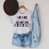I Am A Nice Person Sublimation Transfer, casual outfit with a printed t-shirt, denim shorts, a denim shirt, and a brown hat hanging on a wall.