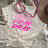 Stacked Pray Pink Screen Print Transfer, a beige t-shirt with pink lettering and prayer phrase print, dressed with pink beaded necklaces, placed on a multicolored woven rug with white crochet decor and a beige hat.